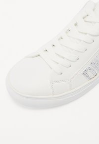 White sneaker made of smooth synthetic leather, featuring a round toe, flat laces, and rhinestone detailing on the side. Rubber sole texture visible.