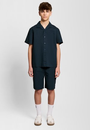 Ragazzo adolescente in piedi che indossa una camicia dark a maniche corte con bottoni, pantaloncini abbinati, calzini bianchi alti e scarpe da ginnastica bianche su uno sfondo semplice.