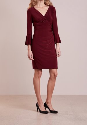 Femme portant une robe bordeaux longueur genou avec des manches trois-quarts cloche et des chaussures à talons noirs, debout sur un sol clair.