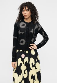 Μαύρο ζακέτα με διάφανο λουλουδάτο σχέδιο, με στρογγυλεμένη λαιμόκοψη, κουμπιά μπροστά και μακριά μανίκια. Συνδυασμένο με πλισέ, ριγέ φούστα.