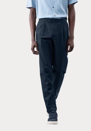 Navy trousers with a straight fit, flat front, and side pockets; paired with a light blue short-sleeve shirt and navy sneakers.