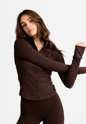 Nuori nainen, jolla on pitkät ruskeat hiukset, pukeutuneena tiukkaan tummanruskeaan vetoketjulliseen takkiin ja legginsseihin, venyttelee käsiään valkoista taustaa vasten.