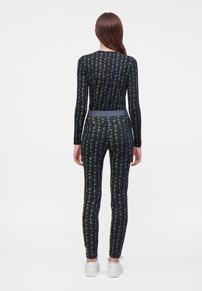 Femme debout de dos portant un haut à manches longues noir à motifs et un pantalon ajusté assorti, avec des baskets blanches sur un fond uni.