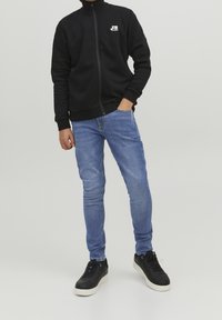 Sudadera negra con cremallera y cuello alto, logo blanco en el pecho; combinada con vaqueros skinny de color azul claro y zapatillas negras con suelas blancas.