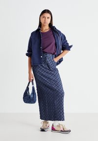 Blå denimjacka över en lila skjorta, kombinerad med en strukturerad maxi-nederdel i marinblått. Accessoarer inkluderar en liten blå handväska och beiga sneakers.