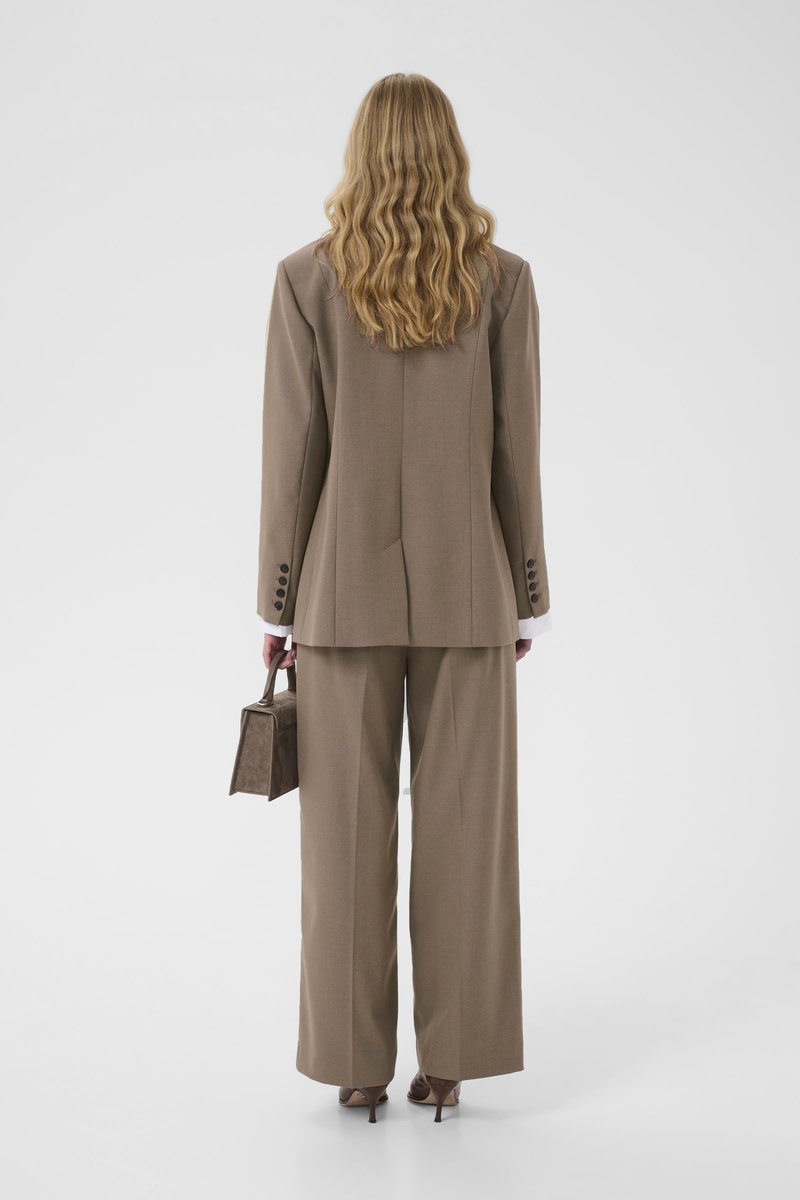 Costume marron sur mesure avec pantalon à jambes larges et blazer long, détails comprenant des manches boutonnées et un petit sac à main marron. Fond uni.