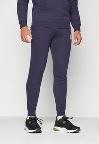 Marineblauwe sweatpants met een slim fit en geribde manchetten. Voorzien van een gouden logo-accent op de dij. Gecombineerd met zwarte sportschoenen.