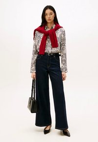 Woman wearing paisley shirt, red sweater draped over shoulders, wide-leg dark jeans, black heels, and holding a black handbag against white background.