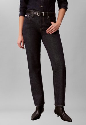 Person iført mørk denimjeans, sort bælte, sort langærmet skjorte og sorte ankelstøvler med hæl, stående mod en grå baggrund.