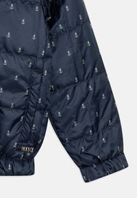 Jachetă puffer marină dintr-un material sintetic neted, cu un model de ancore în întregul său, de culoare gri deschis, având manșete elastice și eticheta brand-ului.
