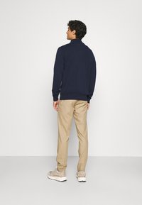 Mørkeblå strikket sweater med høj krave, parret med lyse beige bukser og beige sneakers, set bagfra mod en ensfarvet baggrund.
