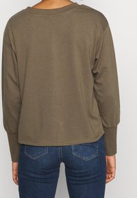Top de manga larga en verde oliva con un corte holgado y un escote amplio, combinado con unos pantalones vaqueros de mezclilla azul oscuro que cuentan con bolsillos traseros y detalles de costura.