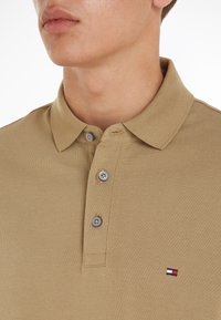 Μπεζ πόλο πουκάμισο από υφή ύφασμα, με γιακά, τρία γκρι κουμπιά και ένα μικρό κεντημένο λογότυπο με κόκκινες, λευκές και μπλε λεπτομέρειες.