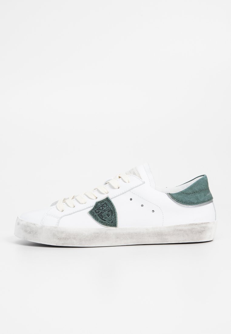 Sneaker bianco a basso profilo con tallone e emblema in suede verde, lacci bianchi e suola leggermente usurata e sporca su sfondo bianco.