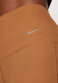 Nike leggings meleg barna árnyalatban, bordás textúrával, magas derékkal és kontrasztos színű logóval.