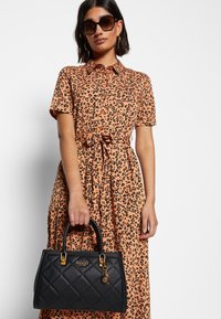 Sac à main noir matelassé avec garniture dorée, tenu dans une main. Le modèle porte une robe courte à manches courtes imprimé léopard orange avec un détail de taille nouée.