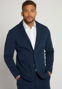 Blazer blu navy realizzato in un tessuto morbido, con revers a punta, due tasche frontali e due bottoni. Indossato sopra una camicia bianca.