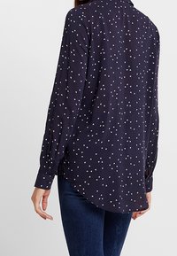 Blusa a manica lunga blu navy con piccoli motivi a triangolo bianchi sparsi, indossata con jeans blu scuro, vista da dietro.