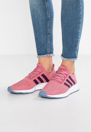 Person wearing pink knit sneakers with dark stripes and blue-gray soles, paired with frayed hem blue skinny jeans on white floor.