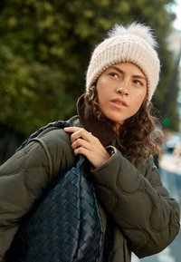 Knit beanie in soft beige with a fluffy pom-pom; green quilted jacket; black woven shoulder bag with textured detailing.