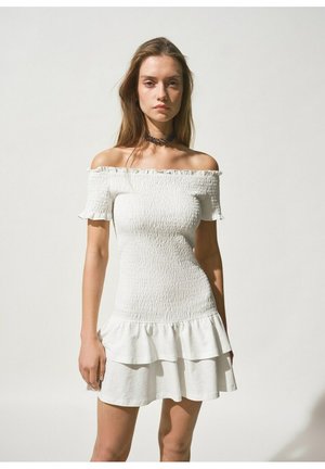 Jeune femme portant une robe blanche smockée à épaules dénudées avec un ourlet à volants et un collier ras-du-cou sombre, debout devant un fond clair uni.