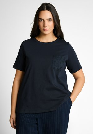 Femme aux longs cheveux bruns portant un chemisier noir à manches courtes avec une poche brodée et un pantalon à fines rayures foncées, debout avec une main dans la poche.