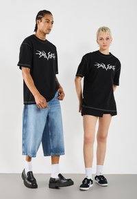 Black oversized t-shirts with white graphic designs, paired with light blue wide denim shorts and dark shoes, set against a plain background.