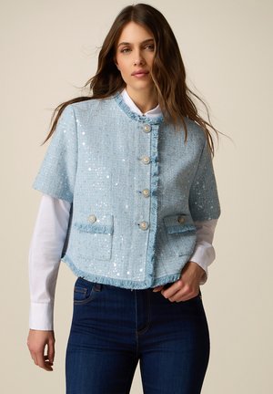 Donna che indossa una giacca corta in tessuto azzurro chiaro con bordo a frange e bottoni sopra una camicia bianca a maniche lunghe e jeans blu scuro.