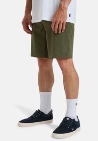 Pantaloni corti verde oliva realizzati in tessuto morbido, con una piccola patch con logo sull'orlo. Indossati con calzini bianchi e sneakers scure in camoscio.