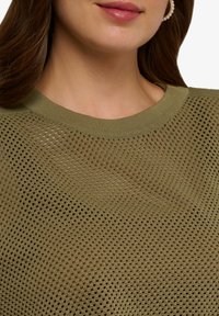 Olivově zelený pletený svetr s výstřihem do vydlabaného tvaru a otevřeným síťovaným vzorem, který ukazuje lehkou a prodyšnou strukturu. Ozdobený žebrovaným výstřihem.