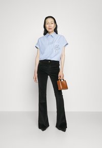 Light blue striped short-sleeve shirt with a side graphic, paired with black flared corduroy pants. Holding a small brown handbag.