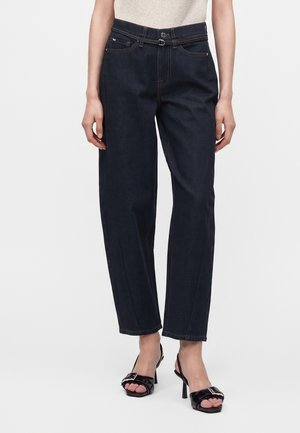 Frau trägt dunkelblaue Jeans mit geradem Bein, einem kleinen schwarzen Gürtel und schwarzen offenen Sandalen mit hohen Absätzen.