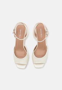 Weiße offene High Heels mit glänzender Oberfläche, quadratischer Vorderseite und einem Knöchelriemen mit silberner Schnalle. Flache Sohle mit gepolstertem Innenbereich.