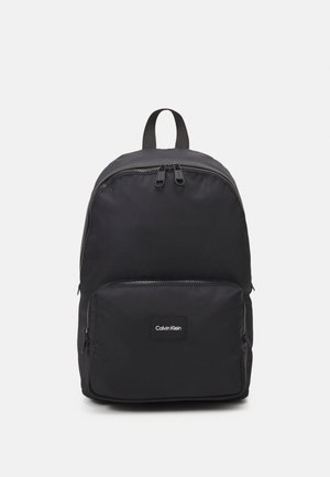 Zaino in nylon nero con doppi zip, una tasca frontale e una toppa logo rettangolare. Presenta spallacci imbottiti e una parte superiore arrotondata.