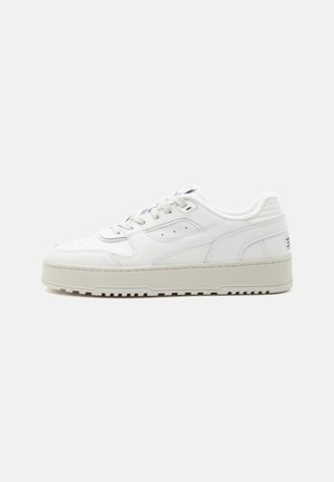 Weißer Sneaker in niedriger Ausführung mit dicker Sohle, Schnürverschluss vorne, perforierten Details und gepolstertem Schaft auf schlichtem hellgrauem Hintergrund.