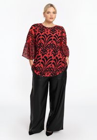 Rote Bluse mit schwarzem Blumenmuster, runder Halsauschnitt und Glockenärmeln, kombiniert mit schwarzen, weiten Lederhosen und spitz zulaufenden Schuhen.
