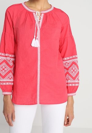Rotes langärmliges Bluse mit weißen, gestickten geometrischen Mustern auf den Ärmeln und einem gebundenen Tassel-Ausschnitt, getragen zu weißen Hosen.