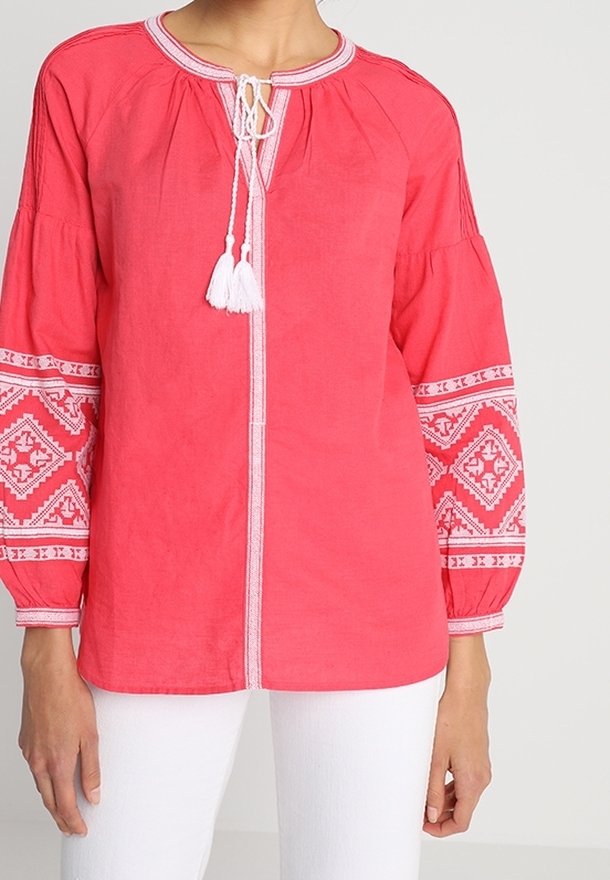 Blouse rouge à manches longues avec des motifs géométriques brodés blancs sur les manches et un col à pompons noué, portée avec un pantalon blanc.