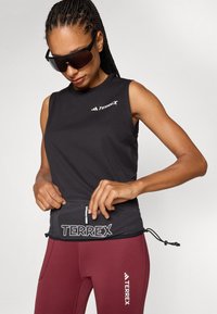 Sort, ermeløs treningsgenser med elastisk nederkant, med "TERREX"-logo, parret med vinrøde leggings. Glatt, lett stoff.