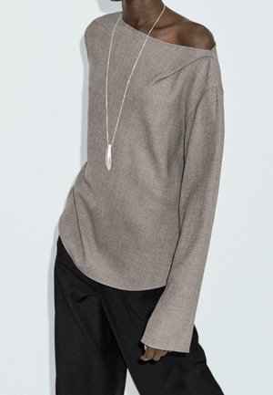 Person wearing a taupe off-shoulder long-sleeve top, black pants, and a long silver necklace with a pendant.