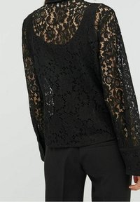 Zwarte bloemenkant lange mouw top met doorschijnende delen, gedragen over zwarte broek, die gedetailleerde kantpatronen op de rug en mouwen toont.