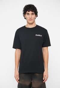 Jovem com cabelo escuro e encaracolado, vestindo uma t-shirt preta com o logotipo "Double A" e calções cargo escuros, de pé contra um fundo branco liso.