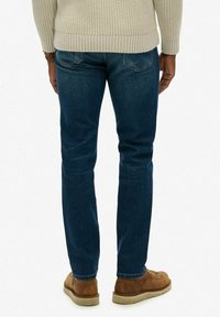 Persona vestida con jeans azul oscuro, suéter de punto beige y zapatos de cuero marrón, de pie con la espalda hacia la cámara.