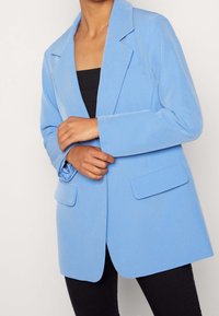 Lichtblauwe blazer van textiel met een gepaste pasvorm, voorzien van notch-uitsparingen en twee voorkruiszakken. Gekozen over een zwart top.