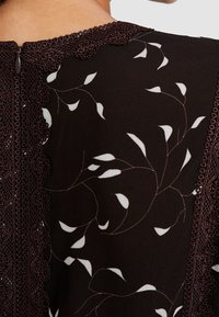Top marrone scuro con un motivo floreale in bianco e beige. Presenta un bordo in pizzo testurizzato lungo il collo e una chiusura con zip sul retro.