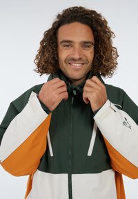 Herenjacket in groen, oranje en wit; voorzien van een ritssluiting aan de voorkant, verstelbare kraag, elastische manchetten en twee zijzakken. Waterdicht materiaal.