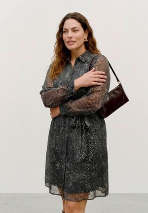 Une femme porte une robe noire à manches transparentes et à motifs, des boucles d’oreilles dorées et un sac à bandoulière foncé, sur un fond uni.