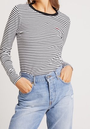 Kvinde iført sort-hvid stribet langærmet skjorte med sort krave og lyseblå højtaljede jeans, hænderne i lommerne.