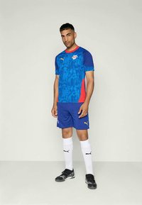 Muž stojí oblečený v modrej a červenej vzorovanej futbalovej dres, modrých šortkách, bielych ponožkách Puma a čiernych kopačkách na jednoduchom pozadí.