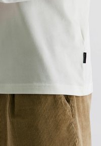 Witte T-shirtzoom met zwart Pierre Cardin-label aan de zijkant, gecombineerd met lichtbruine corduroy broek met zichtbare zak en verticale ribbels.